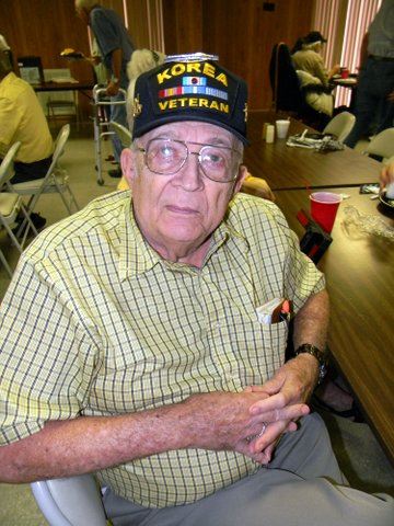 Man sitting, with hands folded, wearing a checked shirt and Korean War veteran hat