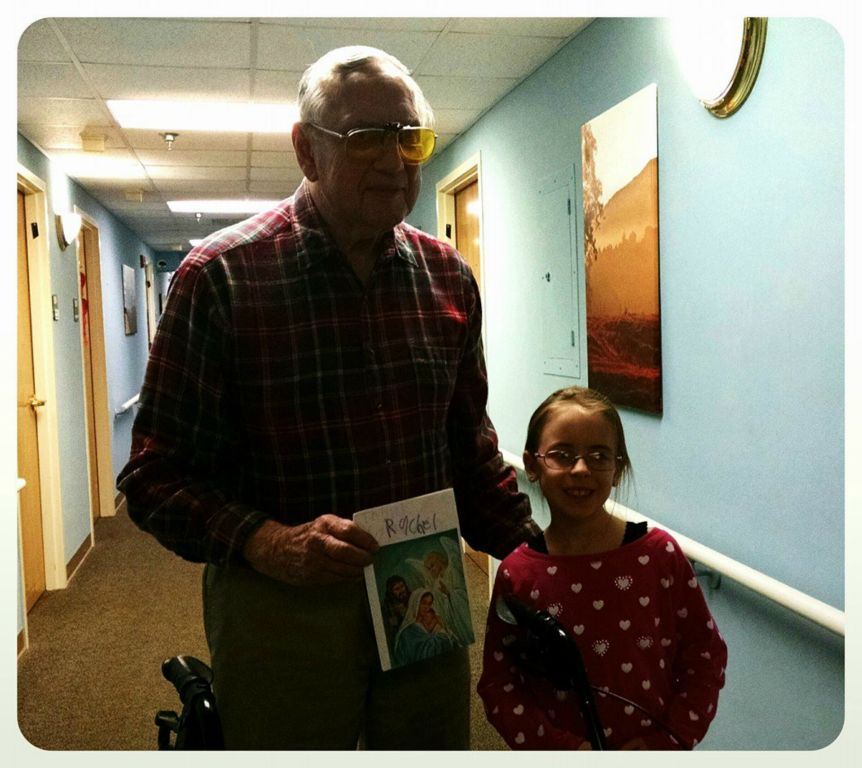 Man in red plaid holds up card with angels on it and the name Rachel, beside girl in red heart sweater