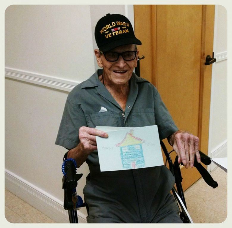 Man sitting in walker wearing glasses and World War II Veteran hat, holds up card with house drawn on it