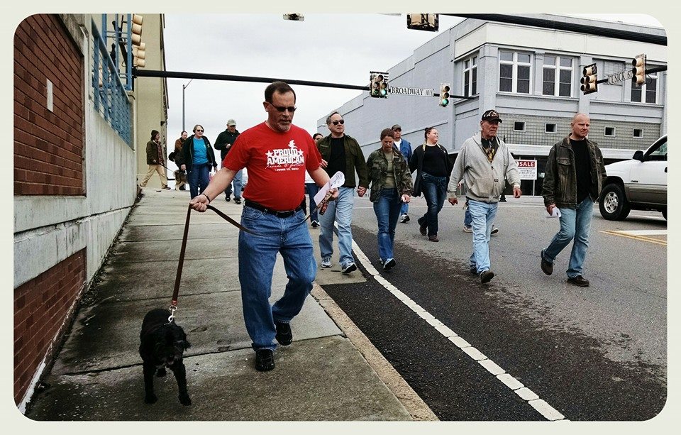 Man in red Proud american shirt walks black dog down street with Awareness walkers