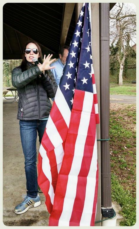 Woman with microphone stands with hand pointing to American flag
