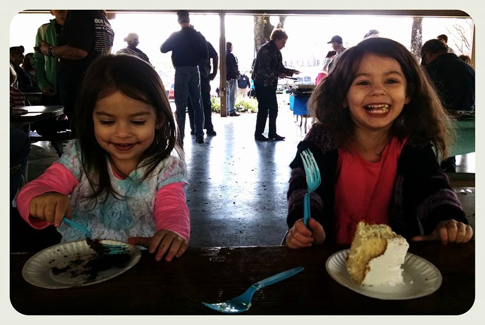 Two young girls sit beside each other enjoying cake at a picnic table