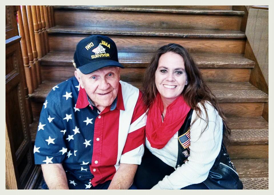 Man wearing Iwo Jima Survivor hat and American flag shirt smiles, sitting on wooden steps with smiling yong woman