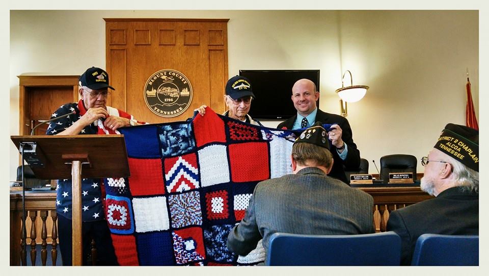 Man wearing Iwo Jima Survivor hat and American flag shirt smiles recieving knitted blanket with navy flags