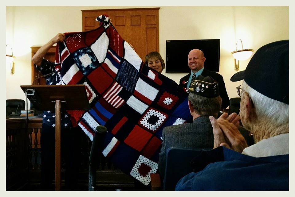 Man wearing Iwo Jima Survivor hat and American flag shirt smiles holding up knitted blanket with navy flags