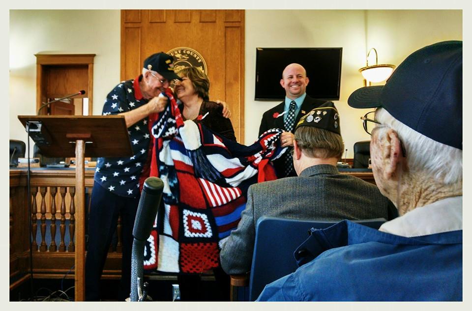 Woman smiles giving knitted blanket with navy flags to man wearing Iwo Jima Survivor hat and American flag shirt