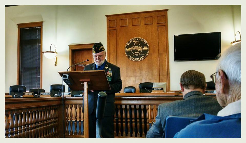 Man in decorated suit and glasses speaks at wooden podium in courtroom