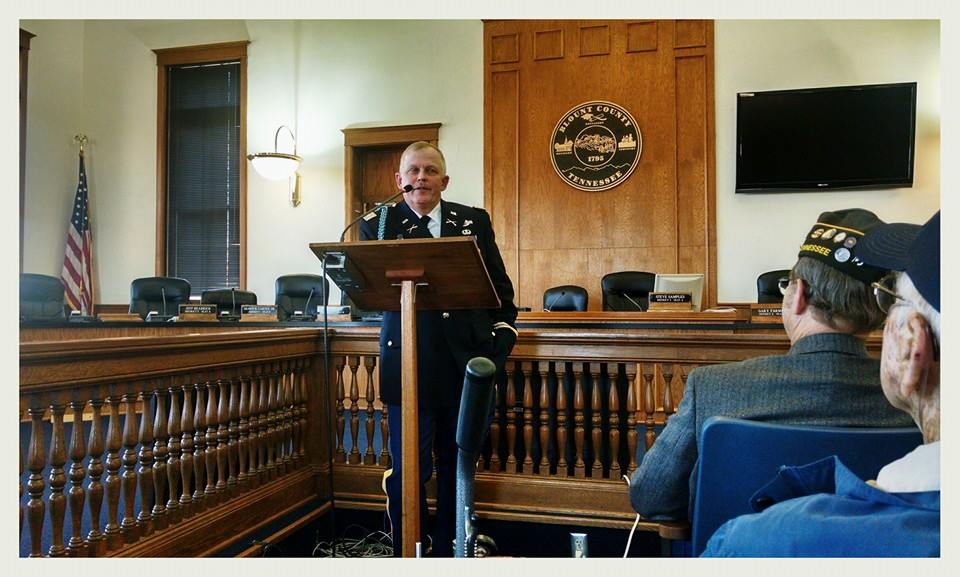 Man in decorated dress uniform speaks at wooden podium in courtroom