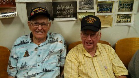 Man with blue shirt and World War II hat sits on couch beside man in yellow shirt and veteran hat