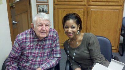 Man in red plaid smiles sitting beside woman in grey shirt, in office