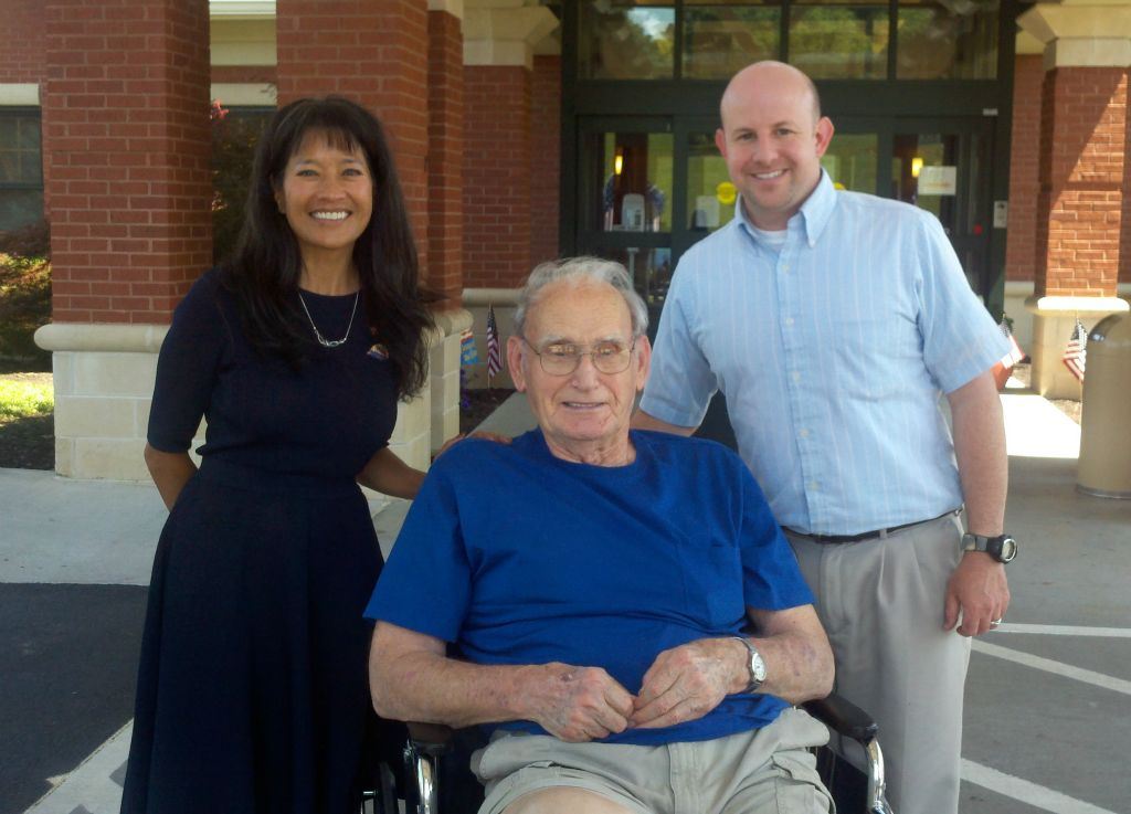 Nathan Weinbaum stands with woman in navy dress behind veteran in blue shirt sittign in wheelchair