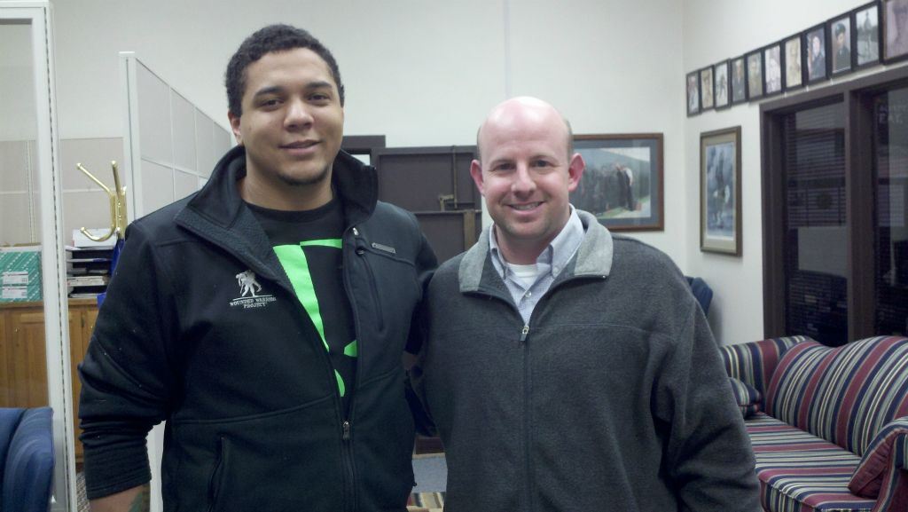 Young man wearing black Wounded Warrior Project sweater side hugs Nathan Weinbaum in grey sweater in office