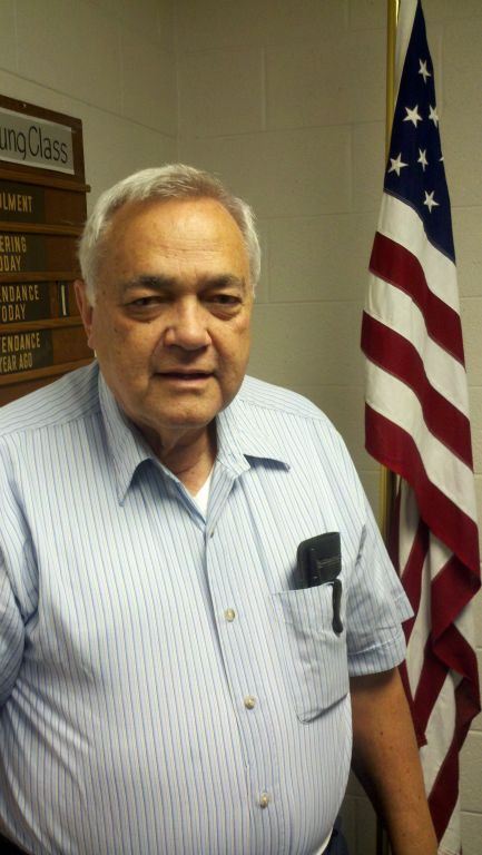 Man in blue striped shirt stands in front of American flag looking into camera