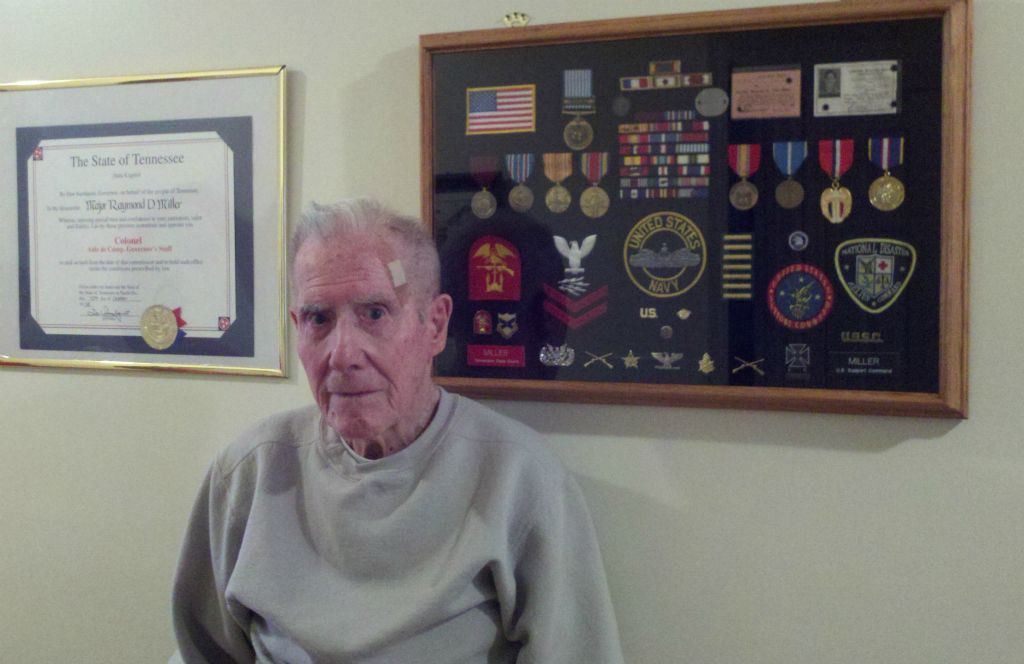 Man in grey sweater stands in front of frame containing medals and pins and patches from Navy