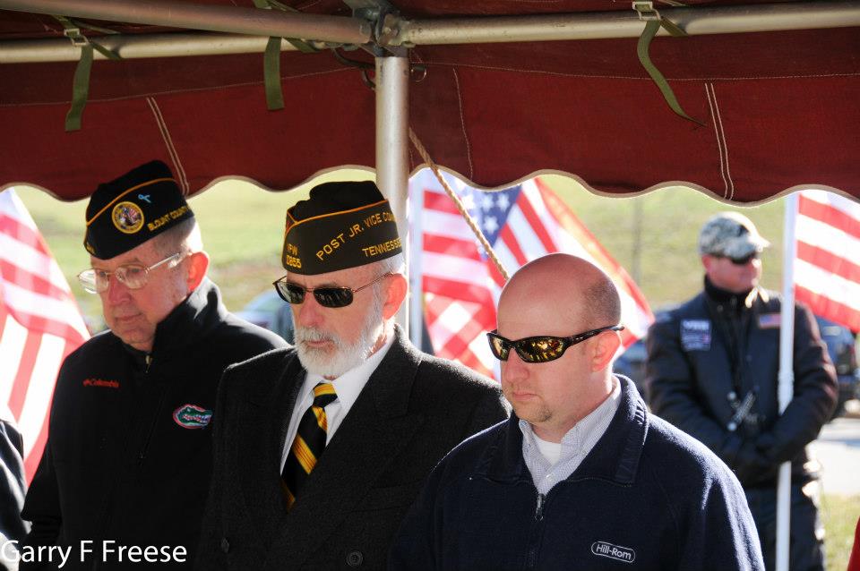 Three men stand under red tent, with American flags in background