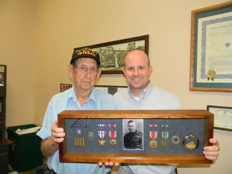 Man wearing World War II hat holds frame with black and white self portrait and medals, with Nathan Weinbaum