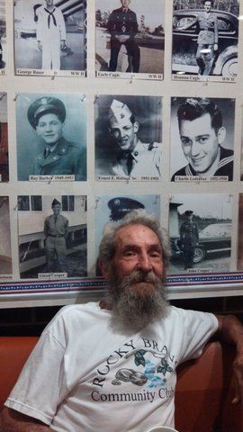Man in white shirt with beard sits on cuoch, with vintage military photogrpahs behind him