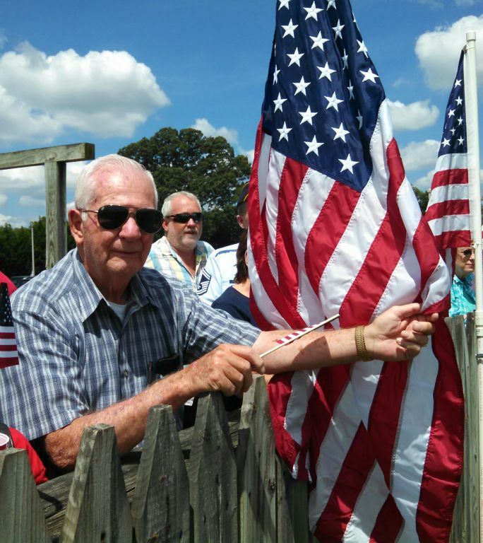 Man in blue plaid and sunglasses leans on wooden fence, holding American flag