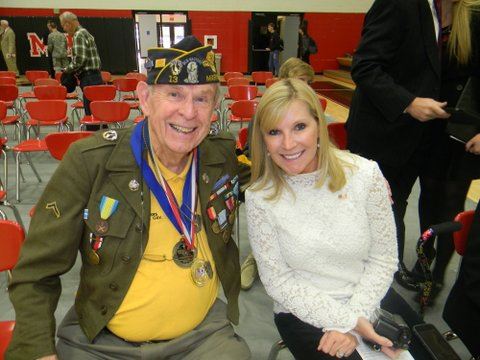 Man wearing yellow shirt, army jacket, cap, and medals smile into camera sitting beside young woman in white shirt