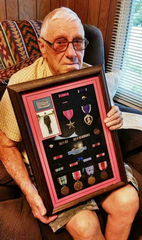 Man with glasses sits in chair, holding frame with self portrait, dog tags, and medals