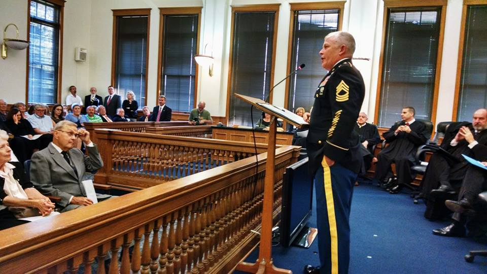 Man in dress uniform stands at podium addressing judges and a court room