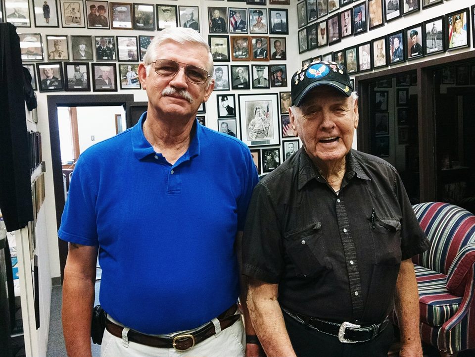 Man with glasses and blue shirt stands beside man in black shirt and veteran hat, both are looking into the camera smiling