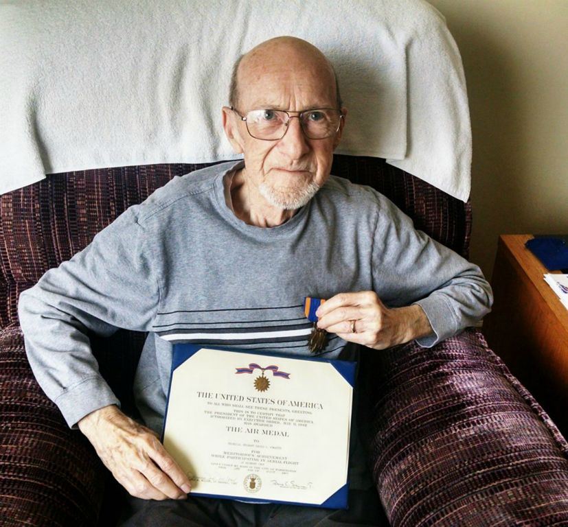 Up close of veteran with glasses, sitting in chair, holding certificate of the Air Medal and also holding the medal to his chest