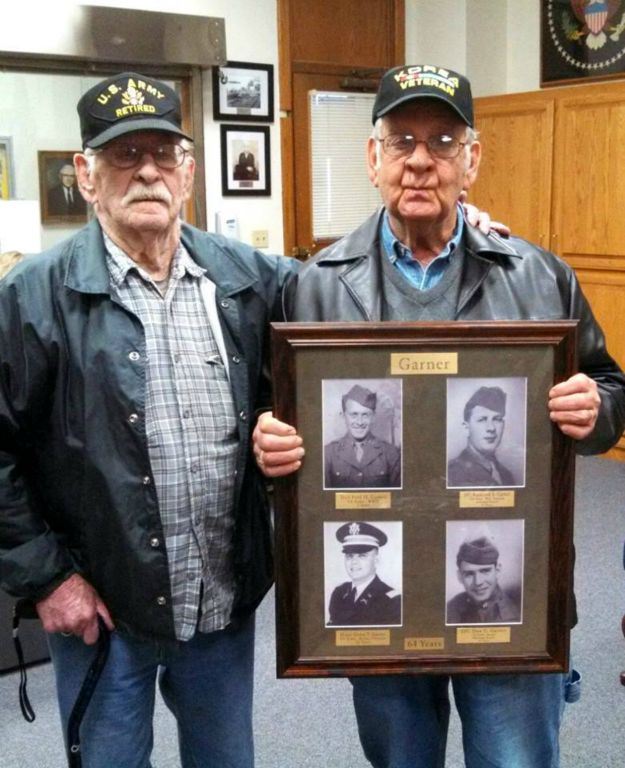 Two men stand together facing camera, one with a US Army Retired hat, and one with a Korea Veteran hat- who is also holding a frame with images of four soldiers with the name Garner