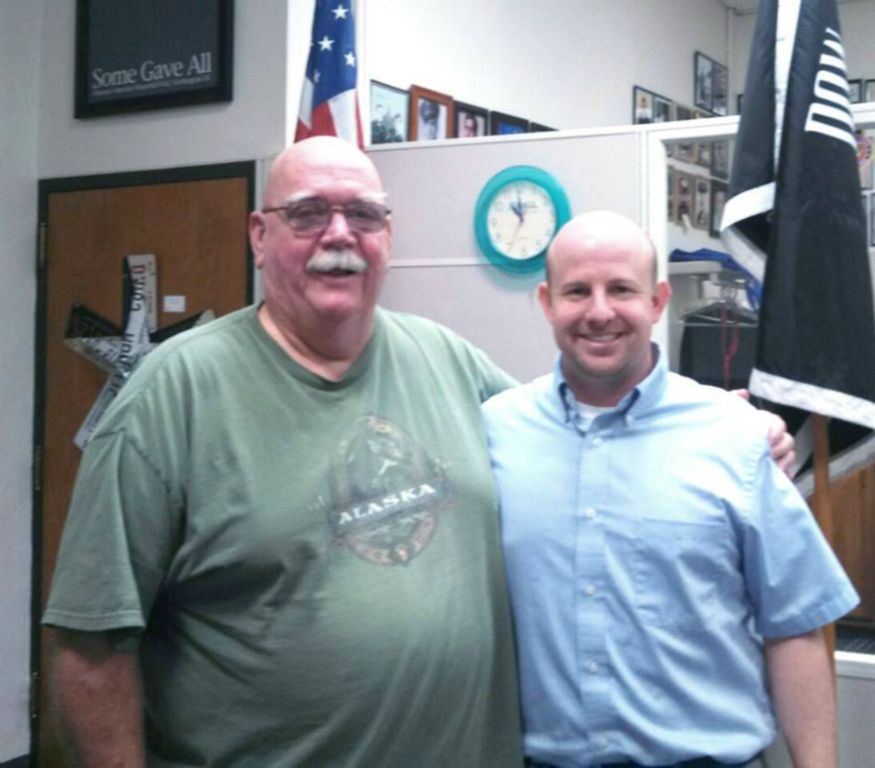 Nathan Weinbaum receives side hug from man with glasses and green shirt, both are looking into the camera smiling