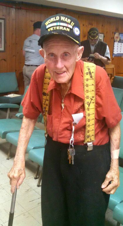 Man wearing orange shirt, yellow suspenders, and World War II Veteran hat, standing an dsmiling into camera