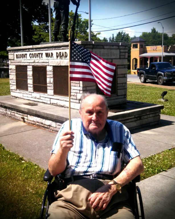 Man in stripe shirt, in wheelchair, holds up American flag in front of Blount COunty War Dead memorial