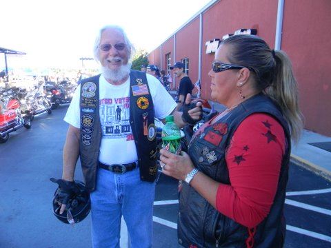 Man in white shirt and black leather vest stands with woman in red shirt and black leather vest in parking lot