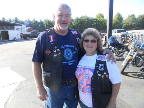 Man drapes arm overwoman in white shirt, standing in parking lot, both are looking into camera smiling