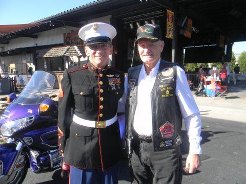 Two men side hug in parking lot in front of purple motorcycle, one in dress Marine uniform and one in biker leather vest