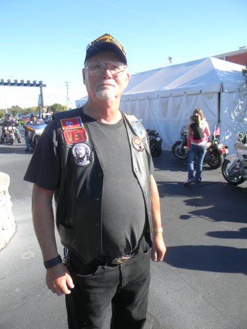 Man in black pants, shirt, and black leather vest, glasses, and black hat stands in parking lot in front of motorcycles