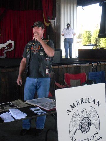 Man in black leather vest and black hat stands behind table with brochures and pamphlets with microphone