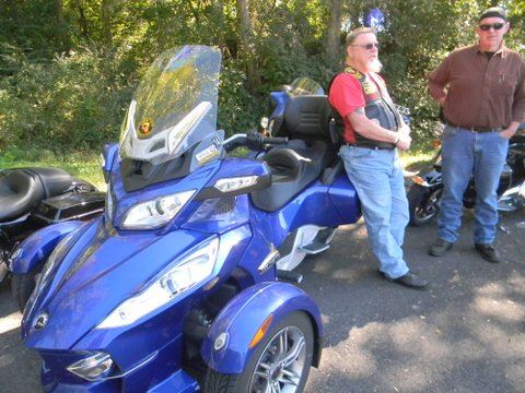 Man leans against blue colored three wheeled motorcycle in parking lot
