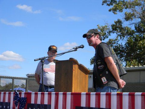 Man wearign hat, sunglasses, and black leather biker vest speaking at podium