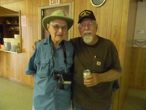 Two men stand together in side hug, one is wearing a camera and the other holds a canned drink, both are smiling into the camera